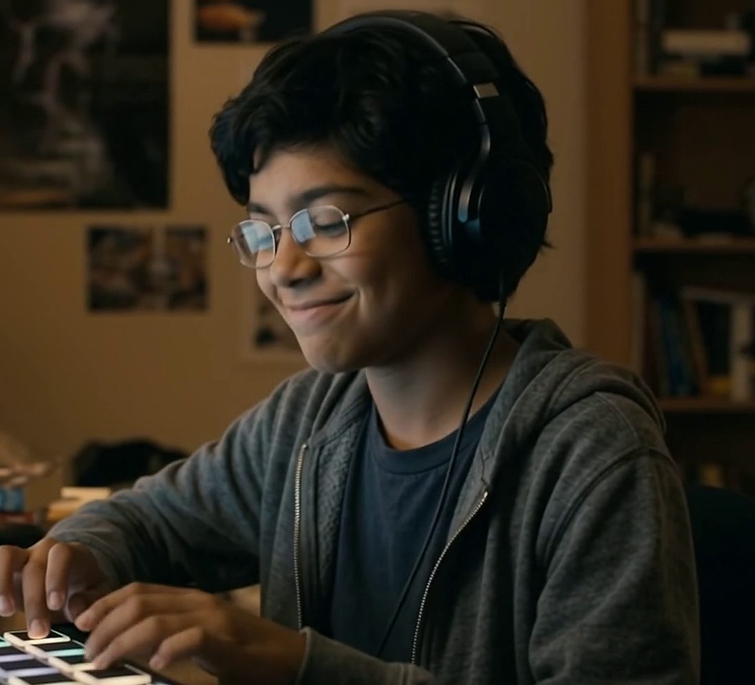 Kaza at his desk at night, wearing big headphones, smiling while he taps a drum pad